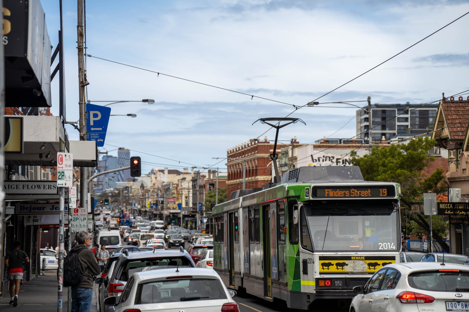 A route 19 tram to Flinders Street in busy traffic on Sydney Road, Brunswick, lined with shops